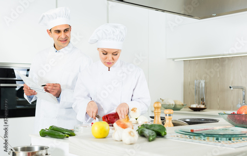 Professional chefs in uniform prepare vegetables with paper recipe on kitchen