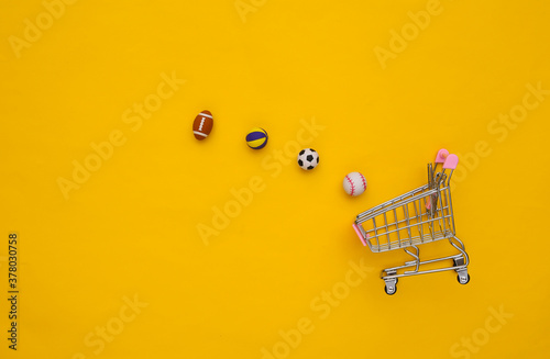 Mini shopping cart with different sport balls on yellow background. Top view