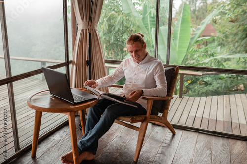 Rear view of a journalist stylish guy read a book in a workplace in loft styled coworking, well dressed, sitting near window with view of garden