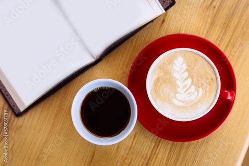 A mug of coffee and a cappuccino coffee mug, an open book next to it.