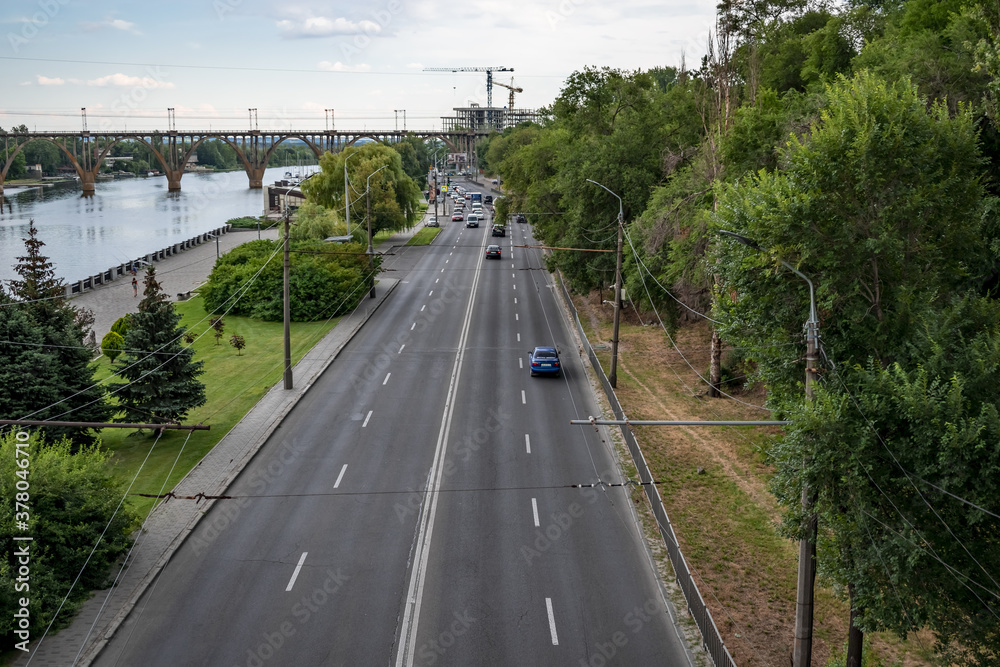 Beautiful city landscape of Sicheslavskaya embankment street in Dnipro (Ukraine). A highway with cars along Dnieper river and the old Merefa-Kherson railway bridge with arched supports on the horizon