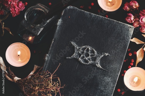 Flat lay of witch's altar with a hand made grimoire with triple moon symbol, and other various items - dried flowers nature elements, candles, ink bottle with black feather next to it on black table