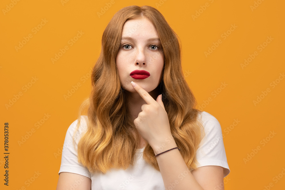 Photo of Pensive young woman with wavy redhead standing with hand raised on chin and looking thoughtfully into a camera Photo of Pensive young woman with wavy redhead standing with hand raised on chin and looking thoughtfully into a camera