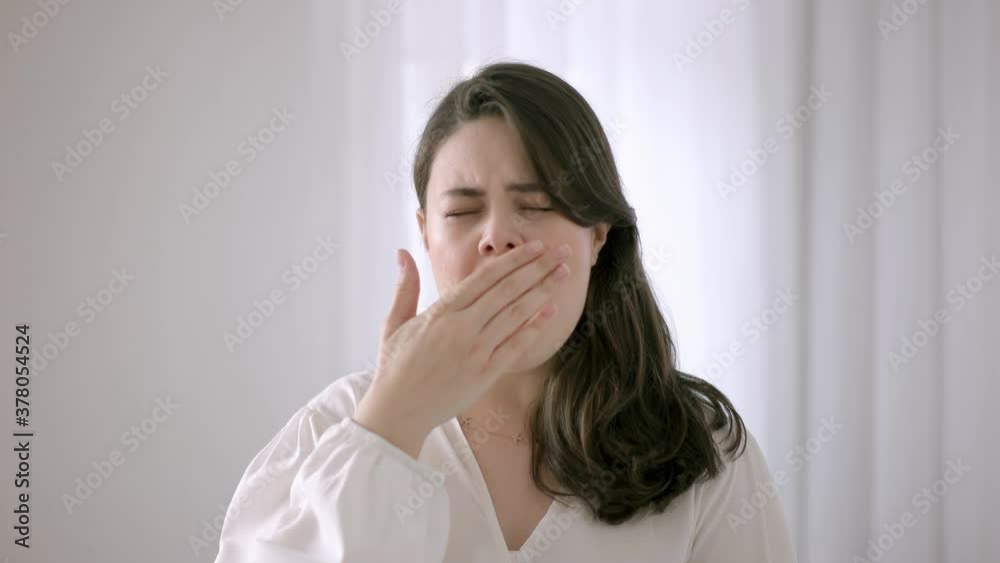 The woman is yawning and covering her mouth with her hand. Woman against white background and looking at camera. The woman is yawning and covering her mouth with her hand. Woman against white background and looking at camera.