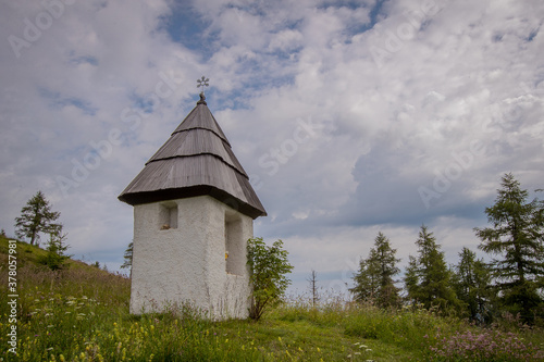 A small white christian chapel on the mountain of Urslja Gora in Slovenia, surrounded by lush greens, flowers and thick white clouds. photo