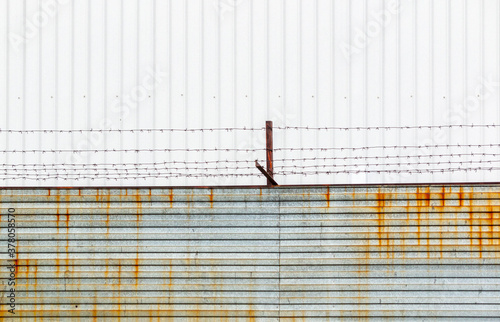 Barbed wire stretched over rusty brackets mounted on a fence made of metal sheets against a cloudy sky.