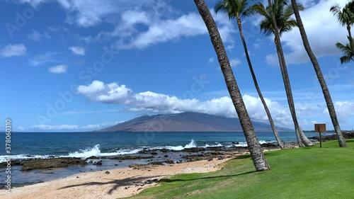 Perfect scene of paradise on Maui, with tall coconut trees in foreground, and golden sand, gentle waves and blue ocean lead to a distant island that is the West Maui Mountains. Paradise in Hawaii.