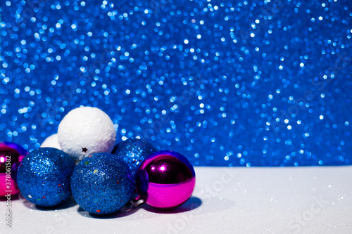 Decorative Christmas balls in blue, white and purple on the left on a white surface. Blue Christmas lights on the background. Side view.Place for inscriptions and greetings