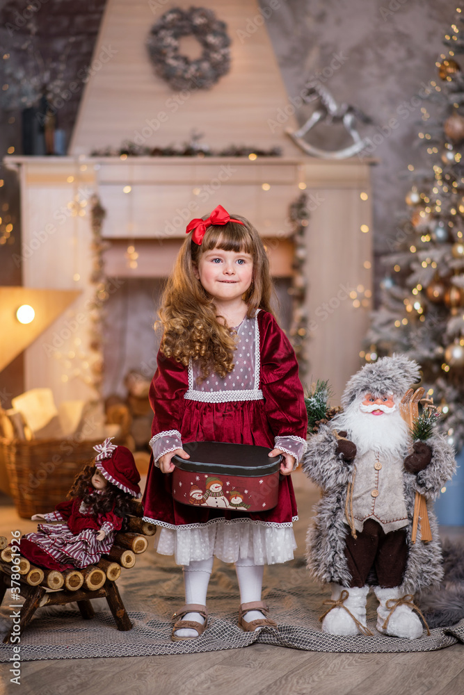 Little beautiful girl in a red dress near the Christmas tree. Girl with a doll are playing Christmas. The child smiles. Beautiful girl with long curly hair. child in santa claus clothes