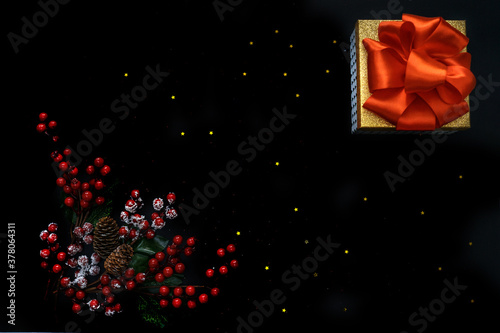 Gift box with red bow and red berries. Black background decorated with stars confetti. Flat lay, top view. Christmas concept.