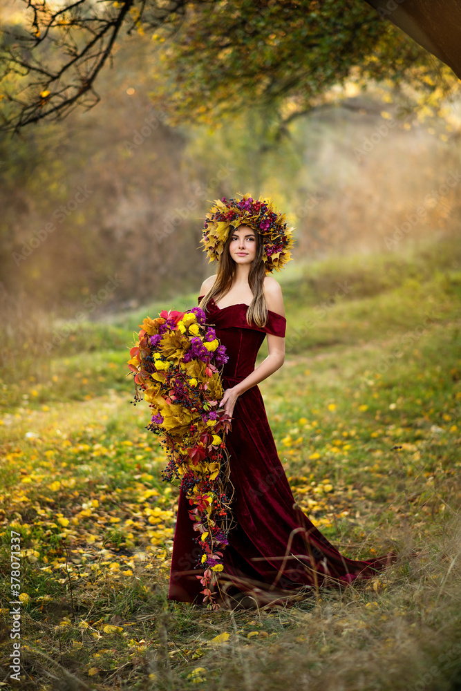A beautiful young woman in an autumn park in a long dress stands under a tree. The girl is holding a composition of flowers and a wreath on her head

