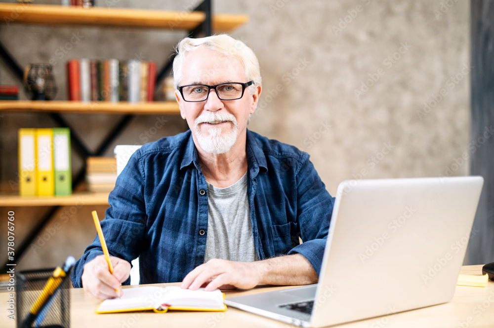 Handsome senior man in smart casual wear is using laptop computer for watching online classes and webinars, he is takes a notes at notebook, while sitting at contemporary home office Handsome senior man in smart casual wear is using laptop computer for watching online classes and webinars, he is takes a notes at notebook, while sitting at contemporary home office