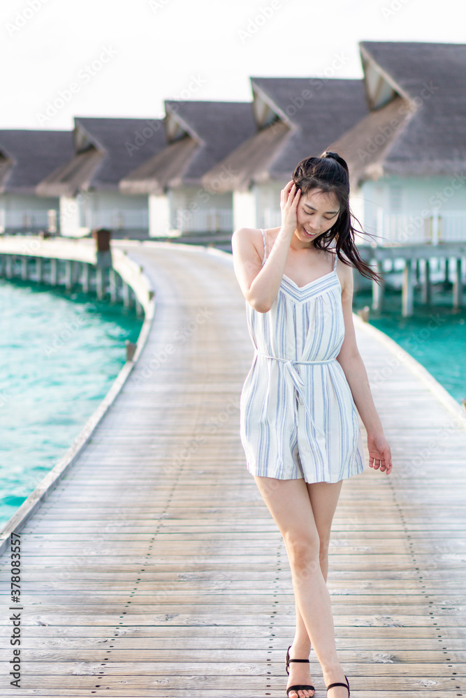 Attractive young woman in white walks over a wooden pier by the sea. Happy young traveller posing on tropical beach enjoys her vacation by sunset sky. Beautiful Woman walking over a wooden jetty.