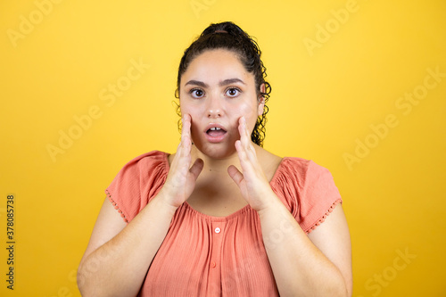 Young beautiful woman with curly hair over isolated yellow background with her hands over her mouth and surprised