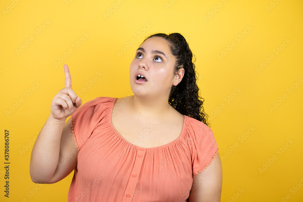 Young beautiful woman with curly hair over isolated yellow background surprised, pointing and looking up.
