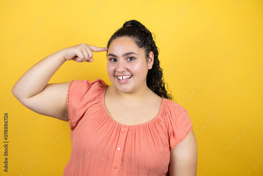 Young beautiful woman with curly hair over isolated yellow background smiling and thinking with her fingers on her head that she has an idea. Young beautiful woman with curly hair over isolated yellow background smiling and thinking with her fingers on her head that she has an idea.