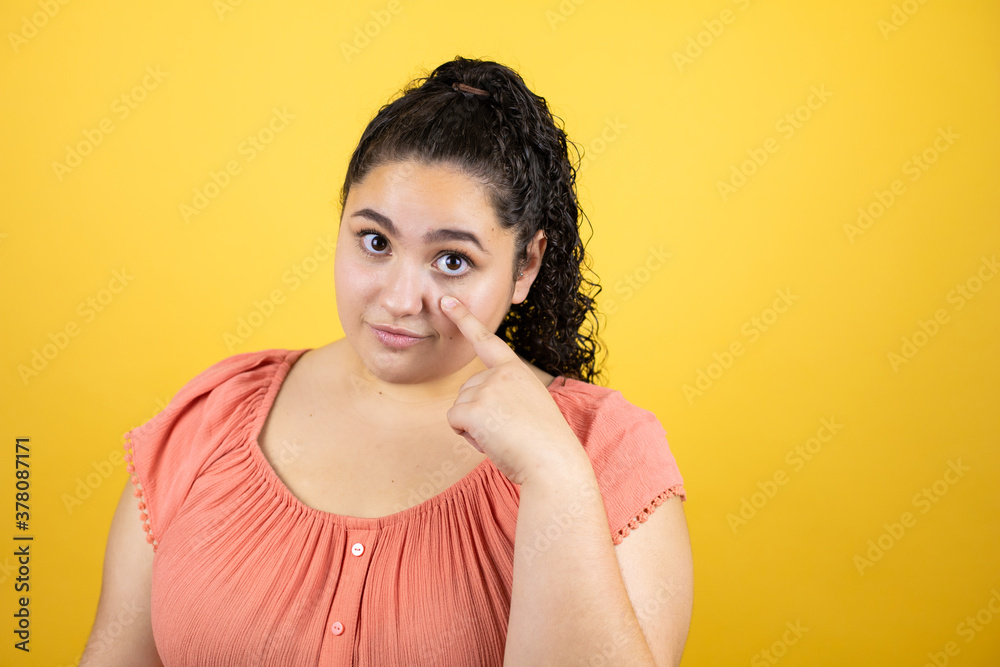 Young beautiful woman with curly hair over isolated yellow background Pointing to the eye watching you gesture, suspicious expression