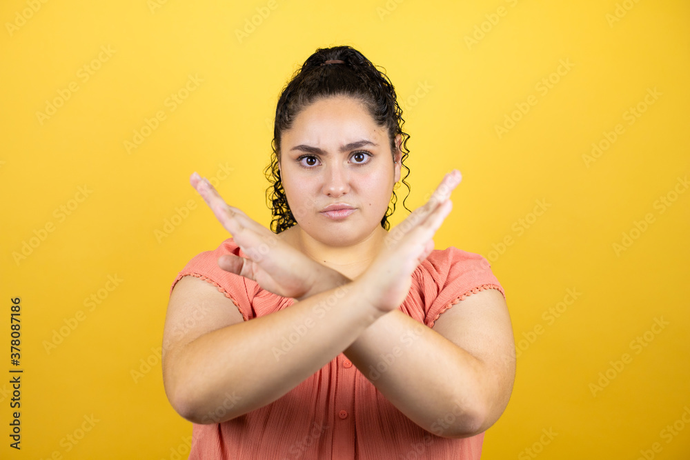 Young beautiful woman with curly hair over isolated yellow background Rejection expression crossing arms doing negative sign, angry face Young beautiful woman with curly hair over isolated yellow background Rejection expression crossing arms doing negative sign, angry face