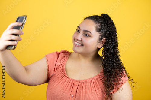 Young beautiful woman with curly hair over isolated yellow background taking a selfie with her phone
