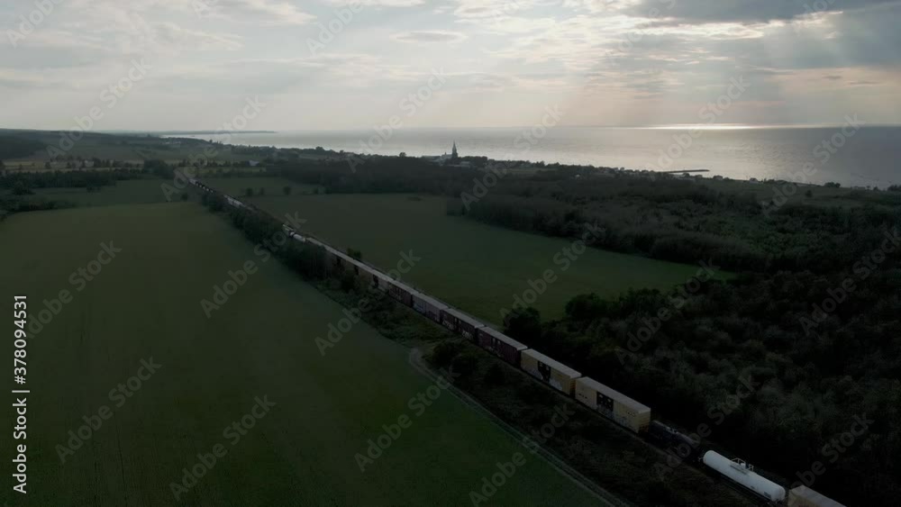 Long Train Passing By On The Railway Through The Green Field In Northern Quebec, Canada. Ray Of Sunlight By The Sea In The Background - aerial drone, ascending shot Long Train Passing By On The Railway Through The Green Field In Northern Quebec, Canada. Ray Of Sunlight By The Sea In The Background - aerial drone, ascending shot