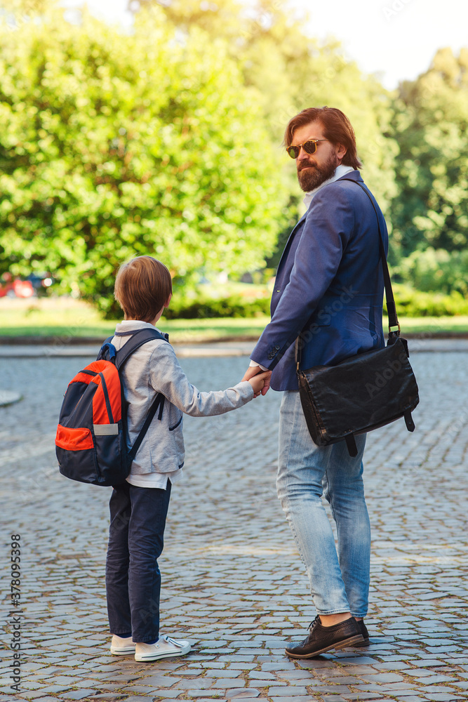 Obraz premium School boy with backpack holding his daddy hand while going to school. Back to school.