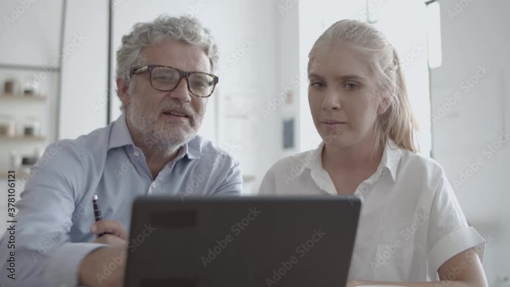 Front view of CEO in glasses talking with female secretary, looking at laptop and holding pen. Successful confident office employees sitting at table and chatting. Business and discussion concept Front view of CEO in glasses talking with female secretary, looking at laptop and holding pen. Successful confident office employees sitting at table and chatting. Business and discussion concept