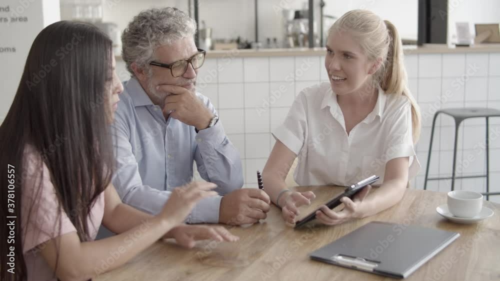 Content colleagues watching tablet screen and sitting at table. Bearded manager holding pen, young businesswomen discussing work in cafe during coffee break. Business and communication concept Content colleagues watching tablet screen and sitting at table. Bearded manager holding pen, young businesswomen discussing work in cafe during coffee break. Business and communication concept