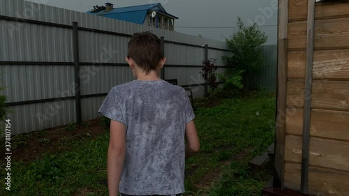 A teenager boy is walking home under heavy rain, the weather is bad and his clothes are wet. Grey cloudy sky and storm in a countryside