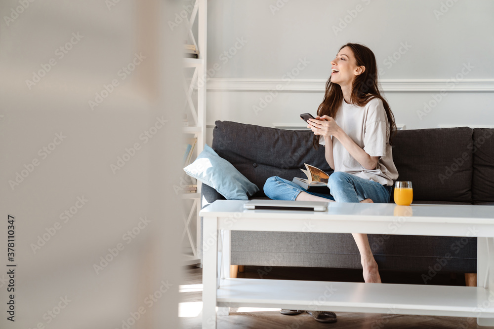 Photo of joyful beautiful girl using cellphone while sitting on couch Photo of joyful beautiful girl using cellphone while sitting on couch