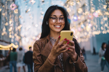 Cheerful millennial blogger enjoying mobile networking during travel vacations for visiting new places, joyful hipster girl in classic glasses for eyes protection reading funny content publication