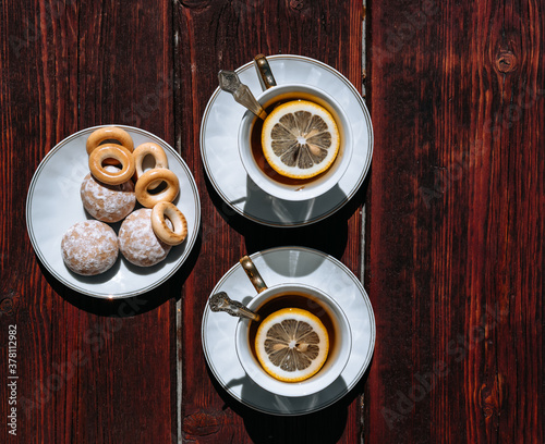 Beautiful white ceramic tableware for tea drinking. Two white cups of black tea with lemon on a wooden table, and delicious bagels and gingerbread in a saucer.