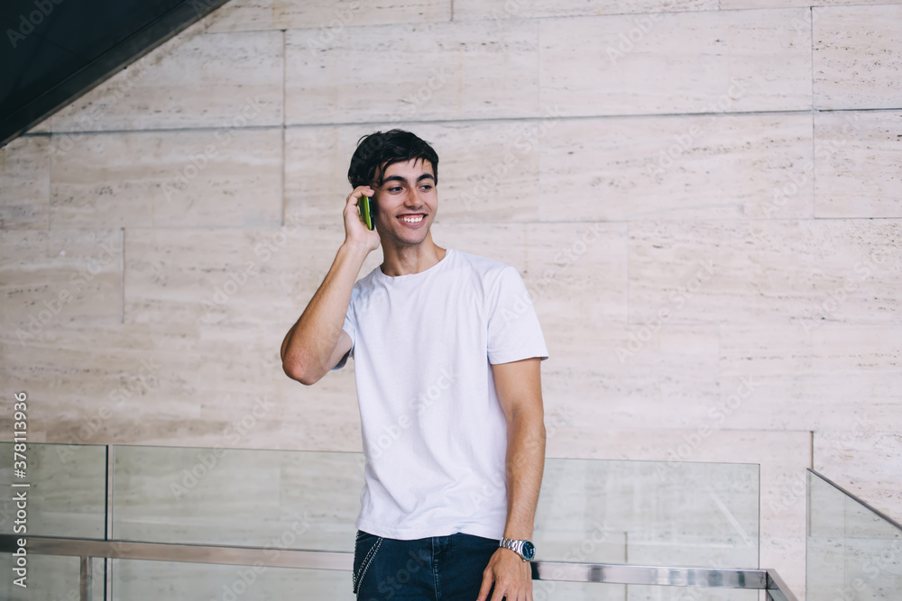 Obraz premium Smiling brunette male in casual wear and white t-shirt talking on mobile phone enjoying connection, handsome 20s positive hipster guy satisfied with smartphone conversation during free time indoors