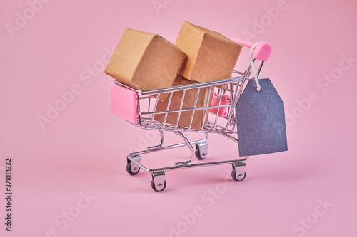 Small shopping cart with blank price tag on a pink background. Shop trolley full of parcels.
