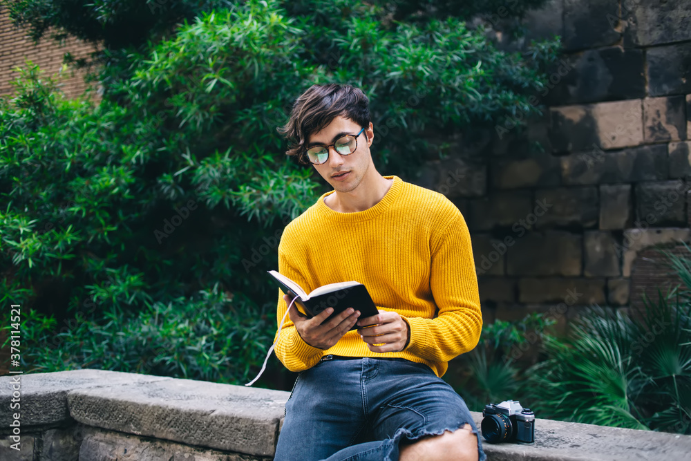 Obraz premium Serious concentrated handsome male reader in eyewear for vision correction enjoying free time on holly and self education, thoughtful hipster guy holding book with interesting plot resting outdoors