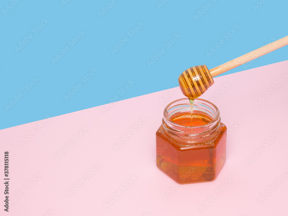 Bee honey drips from a wooden spatula into a glass jar. Pink and blue background. Useful farm products.