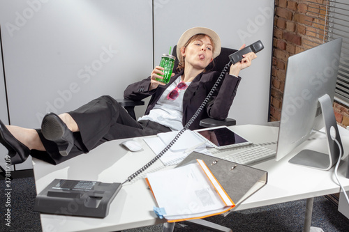 Young female employee at work who stick out her tongue cheekily in direction to the telephone receiver while leaning back relaxed with a refreshing cocktail in her hand, ignoring the phone calls photo