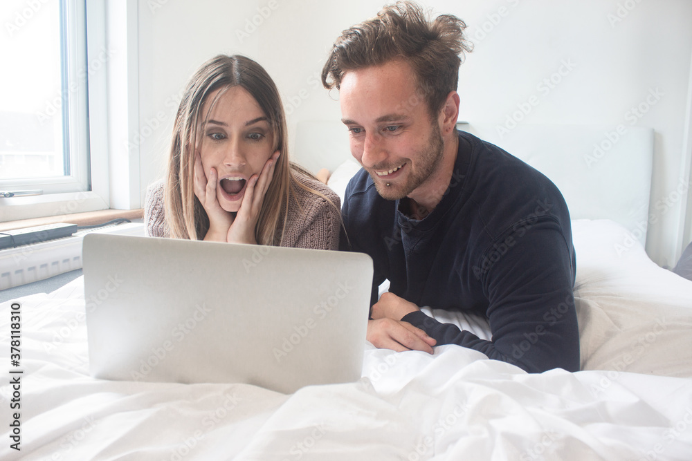 Young couple laying in the bed, looking at the laptop, while the girl is surprised by what she is seeing and the guy laughs. Young couple laying in the bed, looking at the laptop, while the girl is surprised by what she is seeing and the guy laughs.