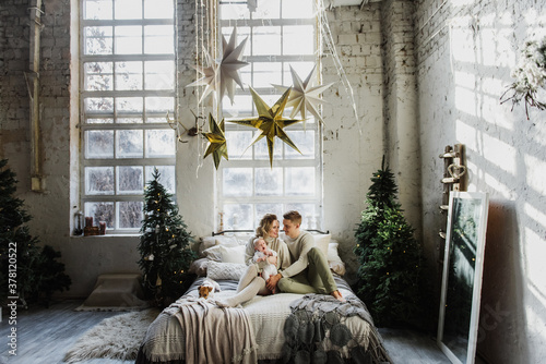 Young women and a man hold a newborn in their arms in the room near the Christmas tree. Family with a newborn and a dog on the bed in a Christmas interior.