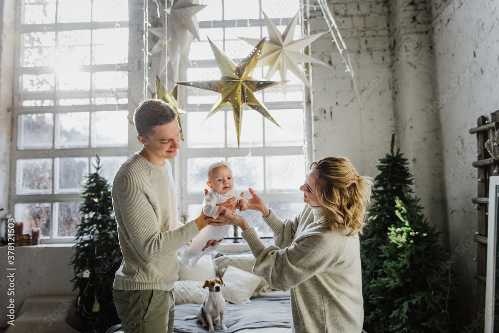 Young women and a man hold a newborn in their arms in the room near the Christmas tree. Family with a newborn in the Christmas interior.