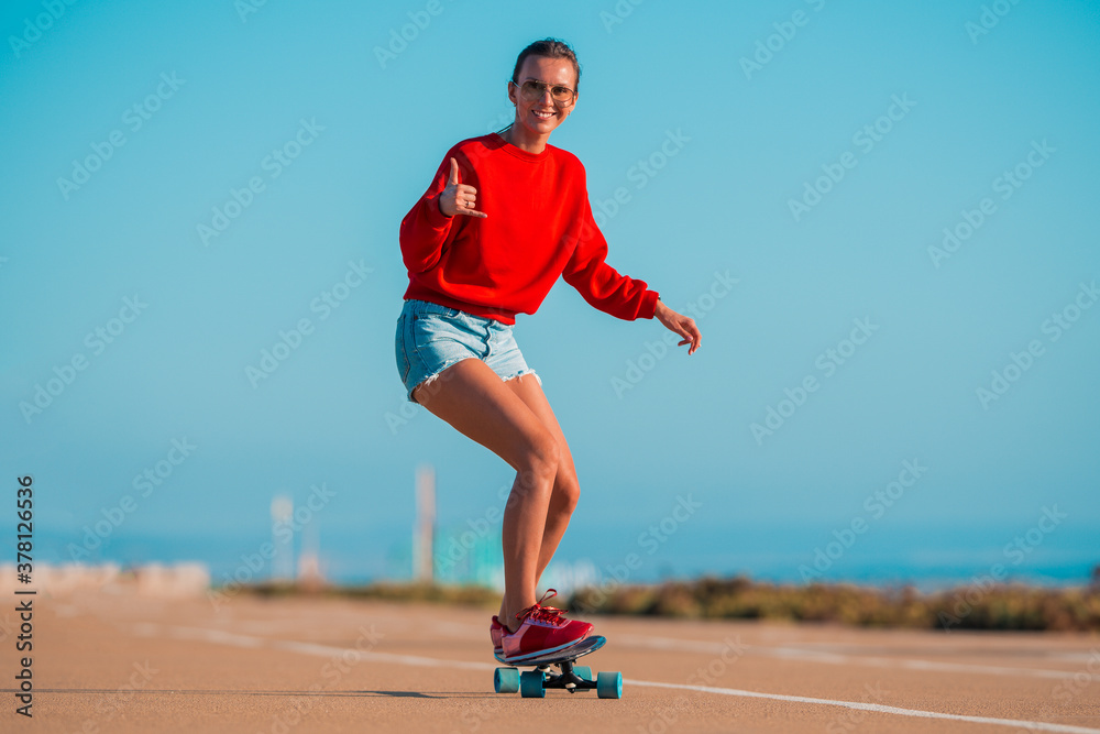 Obraz premium Happy smiling carefree woman in red hoodie and jeans shorts riding surf skateboard and showing shaka sign. Summer fun outdoor activities concept. Street culture.