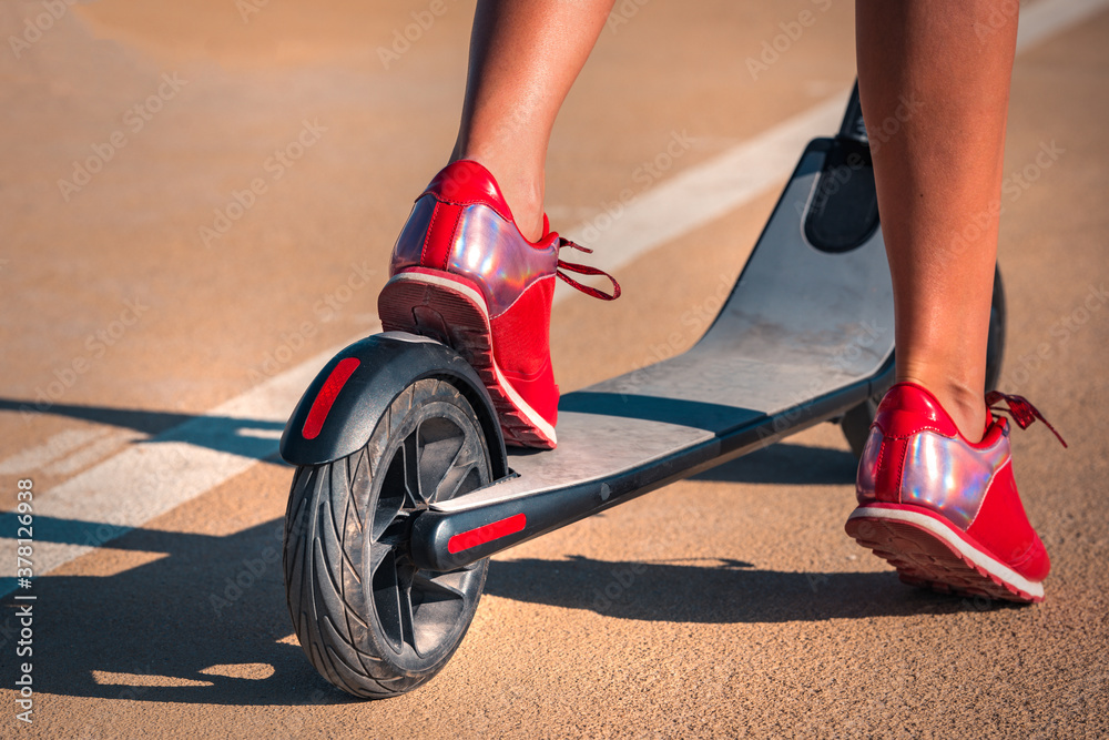 Obraz premium Close up photo of young woman with beautiful legs in red sneakers standing with electric scooter on the road. Summer outdoor activities.