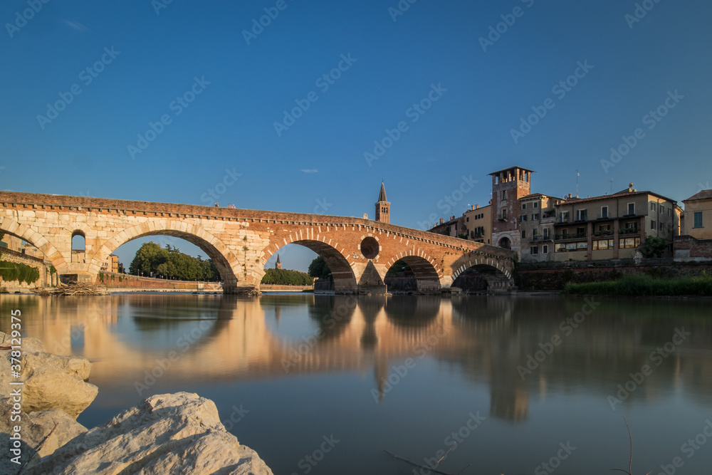 view of the famous Ponte Pietra in Verona, rising above the river of Adige in Italy on early afternoon. Long exposure daylight shot on a sunny evening of an old brick bridge.