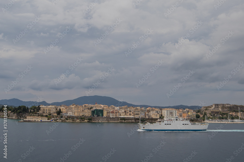 A white ferry boat traveling past the city of Corfu, Greece on a cloudy summer day. View from the sea.