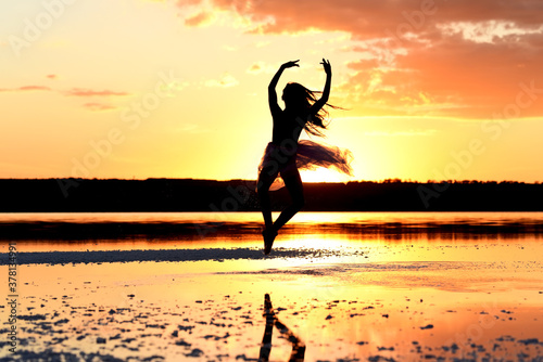 Silhouette of a dancing girl in a ballerina dress against the backdrop of the setting sun on the lake.