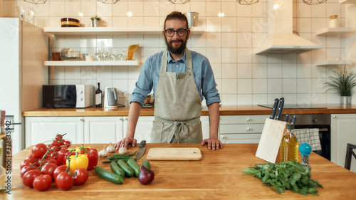 Fotografie Young man, Italian cook in apron smiling at camera while preparing healthy meal with vegetables in the kitchen