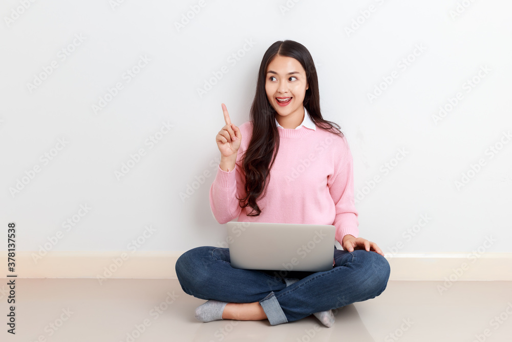 Young attractive Asian woman sitting with laptop on her legs finger pointing up showing copy space with smiley face. Young attractive Asian woman sitting with laptop on her legs finger pointing up showing copy space with smiley face.
