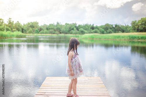 Close-up view of a young Asian girl, lovely, cute wearing a colorful dress, sitting by a pond in the village, closely supervised during school holidays