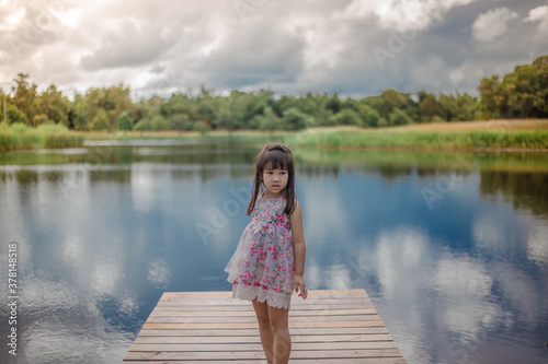 Close-up view of a young Asian girl, lovely, cute wearing a colorful dress, sitting by a pond in the village, closely supervised during school holidays © bangprik