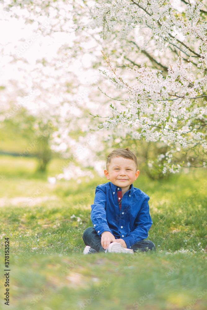 Child boy sits on sunlit glade in blooming garden.
