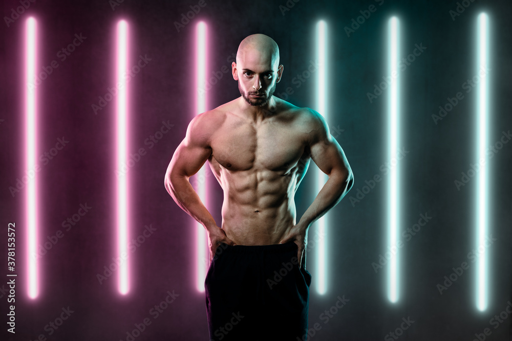 athletic man stand in front of camera look serious straight forward with hands on waistband light up by pink and blue party light tubes in background using the colors symbolize future